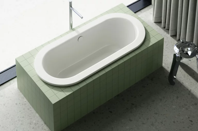 Oval built-in bathtub with green tile surround, freestanding faucet, and curtain by the window.